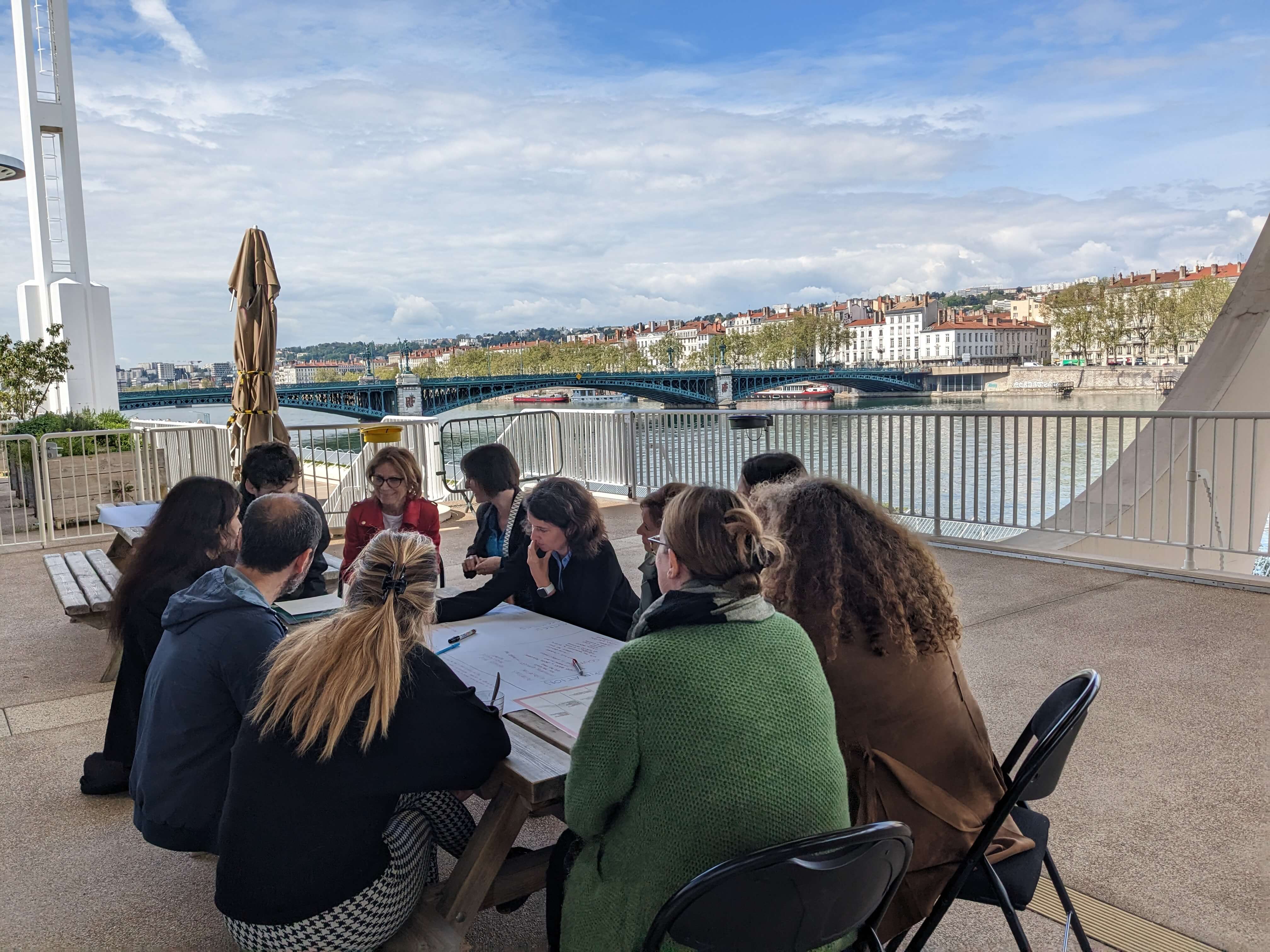 The image shows a group of people sitting on a terrace at a table. The people are engaged in a discussion and are working on a common task.